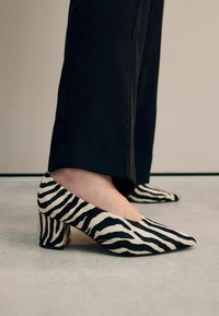 Black and white zebra print high-heeled shoes with a pointed toe and chunky heel, made from textured fabric, positioned near black trousers.