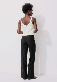 Haut sans manches texturé blanc avec un dos bas, associé à un pantalon large noir et des chaussures à talons noires. Boucles d'oreilles simples visibles.