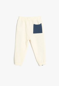 Pantalones jogger de algodón en crema con un detalle de bolsillo azul. Presentan una cinturilla elástica y puños ajustados. Textura suave, diseño casual.