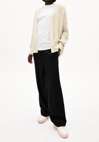 Krémový pletený kardigan přes bílé tričko s kulatým výstřihem, spárovaný s volnými černými kalhotami a bílými teniskami s béžovými akcenty.