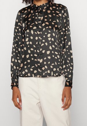 Woman wearing a black satin long-sleeve blouse with beige abstract spots and white high-waisted pants.