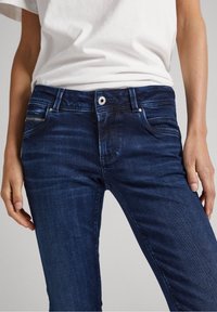 Mörkblå denimjeans med en åtsittande design, som har en framknappstängning, en ficka med dragkedja och subtila slitningar på tyget.