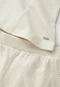 Gros plan sur un tissu doux rayé beige et blanc avec une petite étiquette métallique marquée « NEXT », montrant des coutures cousues et un élastique froncé.