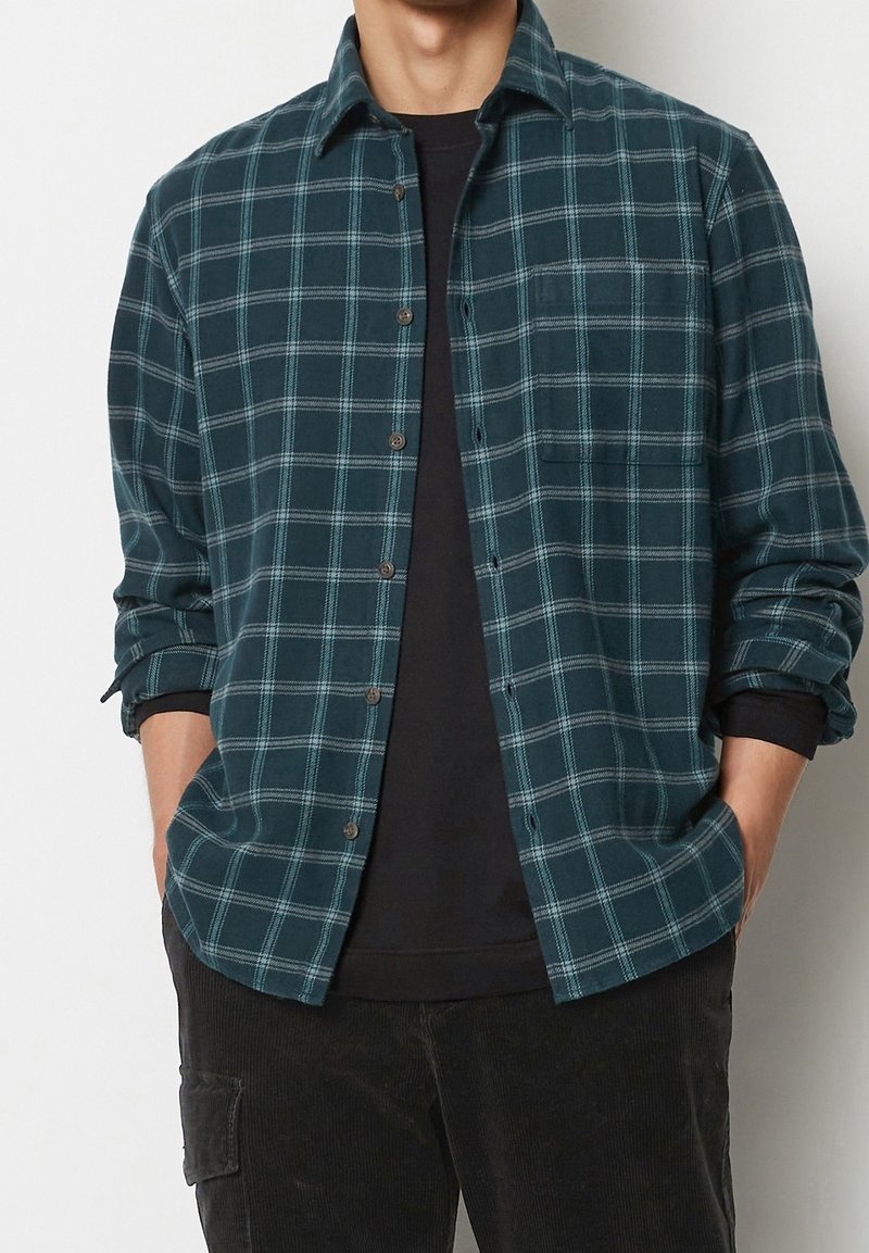 Koszula flanelowa w kratę w ciemnym morskim odcieniu i białym kolorze, z zapięciem na guziki z przodu, długimi rękawami podwiniętymi oraz kieszenią na piersi. Miękka tekstura.