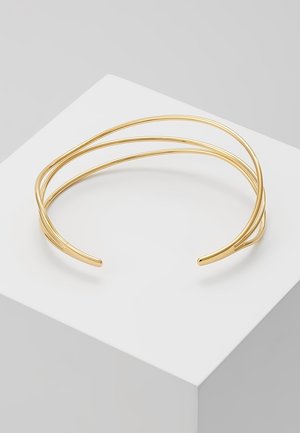 Open armband met drie dunne, in elkaar verweven gouden banden, tentoongesteld op een wit voetstuk tegen een lichtgrijze achtergrond.