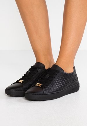 Baskets en cuir noir texturé avec lacets noirs et accents dorés du logo "MK", portées pieds nus.