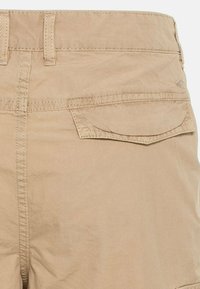 Pantalon beige décontracté avec une poche à rabat à l’arrière et des passants de ceinture visibles, coutures piquées et un léger pli près du bas.