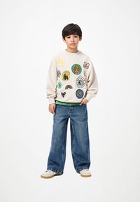 Fant v kremni trenerki s pisanimi obliži, v modrih širokih hlačah in belih športnih copatih, stoji pred enobarvno belo podlago.