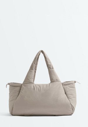 Beige polstret håndtaske med to tykke håndtag og lynlåslommer på siderne på en ensartet lys baggrund.