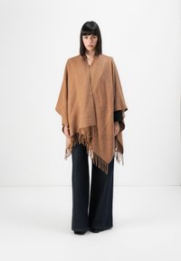 Brun vævet poncho med frynser, der har et tekstureret mønster. Sammen med mørke, flagrende jeans. Glat stof og komfortabel silhuet.