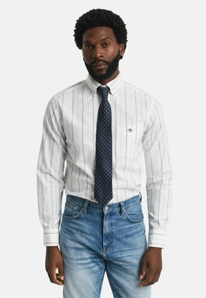 Wit gestreept shirt met opgerolde mouwen, met een kleine borstzak en een marineblauwe stropdas met witte stippen. Gecombineerd met een blauwe denim jeans.
