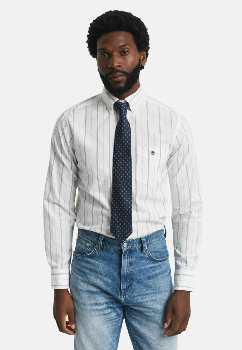 Weiß gestreiftes Hemd mit umgeschlagenen Ärmeln, ausgestattet mit einer kleinen Brusttasche und einer marineblauen Krawatte mit weißen Punkten. Kombiniert mit blauen Jeans.
