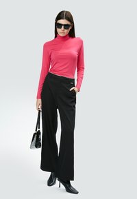 Langärmliger, enganliegender pinkfarbener Rollkragenpullover kombiniert mit hochgeschnittenen, schwarzen, weit geschnittenen Hosen. Zubehör umfasst eine schwarze Handtasche und Sonnenbrille.