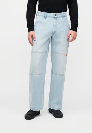 Personne portant un jean droit bleu clair avec des coutures aux genoux et des chaussures noires à enfiler, debout devant un fond blanc uni.