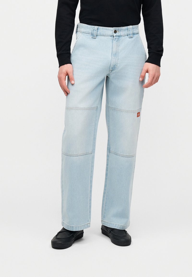 Άτομο που φοράει ανοιχτό μπλε τζιν με ίσια γραμμή ποδιού και ραφές στα γόνατα, μαζί με μαύρα παπούτσια τύπου slip-on, στέκεται μπροστά σε απλό λευκό φόντο.