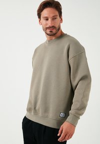 Sudadera caqui hecha de tejido tejido con cuello redondo, hombros caídos y puños acanalados. Presenta un pequeño parche con el logotipo en el dobladillo.