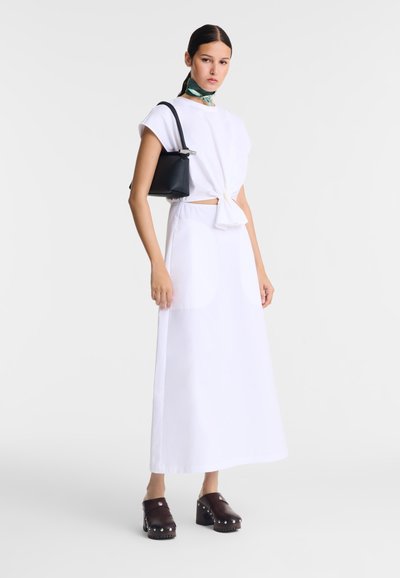 Weißes Kleid mit hohem Kragen, gebundenem Taillengürtel und seitlichem Ausschnitt, kombiniert mit einer schwarzen Handtasche und braunen Clogs mit silbernen Akzenten.