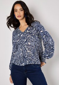 Blaue und weiße gemusterte Bluse mit V-Ausschnitt und langen, puffigen Ärmeln; leichtes Material mit fließender Textur, kombiniert mit dunkelblauen Jeans.