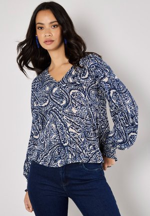 Blå och vit mönstrad blus med V-ringning och långa puffärmar; lättviktsmaterial med en flytande textur, matchad med mörka jeans.
