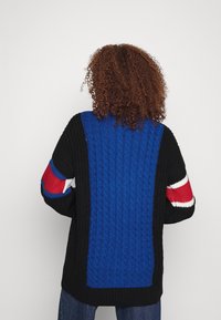 Maglione lavorato a maglia nero con centro a trecce blu e maniche accentuate in rosso. Presenta una vestibilità ampia e un motivo testurizzato.