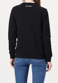 Personne portant un sweat-shirt noir à manches longues Karl Lagerfeld et un jean bleu, vue de dos sur un fond blanc uni.