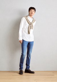Weißes Hemd mit Knöpfen, beiger Pullover über den Schultern drapiert, blaue Jeans und schwarze Schuhe mit goldenen Akzenten. Einfaches, lässiges Outfit.