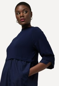 Navyblaues Kleid mit einem strukturierten Oberteil, leicht abgesenkter Taille, kurzen Ärmeln und seitlichen Taschen; glattes Gewebe mit matter Oberfläche.