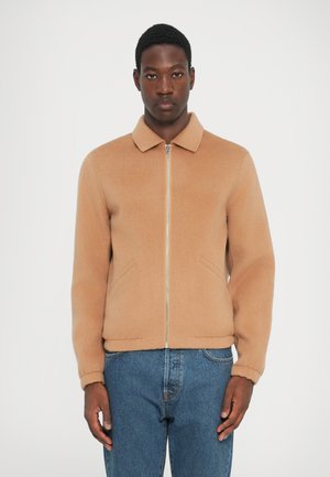 Chaqueta de mezcla de lana marrón con cuello, cremallera frontal y bolsillos laterales. Textura suave con un ajuste relajado. Combinada con jeans azules.