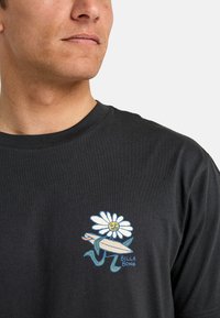 T-shirt en coton noir avec un graphique coloré d'une fleur tenant une planche de surf, avec le texte "BILLA BONG" en bleu en bas.