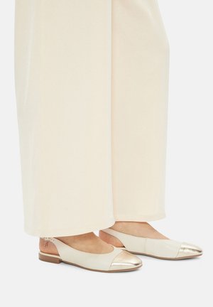 Ballerines couleur crème avec un bout en métal doré, texture en cuir lisse et talon bas et souple, associées à un pantalon large et clair.