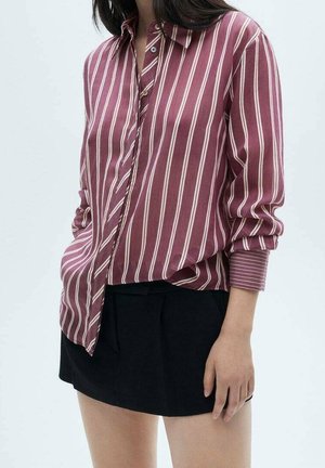 Kvinde iført en maroon og hvid lodret striped skjorte med lange ærmer og sort mininederdel, stående foran en ensartet baggrund.