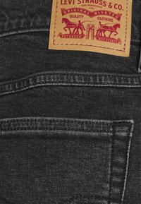 Tela de mezclilla negra con un acabado texturizado, que presenta una etiqueta de cuero marrón con una marca impresa en rojo y un gráfico de un caballo. Costura del bolsillo trasero visible.