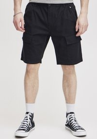 Zwarte cargoshorts gemaakt van stevig materiaal, met meerdere zakken, een elastische tailleband en een comfortabele pasvorm. Draagt met zwarte hoge sneakers.