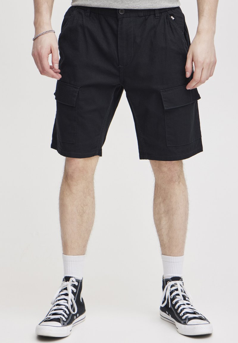 Zwarte cargoshorts gemaakt van stevig materiaal, met meerdere zakken, een elastische tailleband en een comfortabele pasvorm. Draagt met zwarte hoge sneakers.