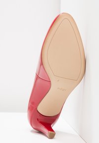 Chaussure à talon haut en cuir verni rouge, avec un bout pointu et un talon aiguille bas. Surface lisse et brillante ; semelle beige avec un marquage minimal.
