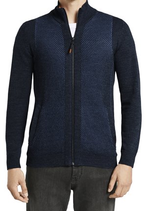Man wearing navy blue textured full-zip sweater with long sleeves and ribbed cuffs, paired with dark pants.