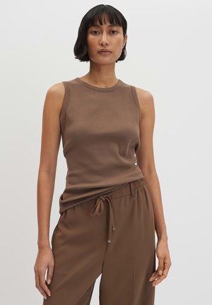 Donna con top marrone a coste senza maniche e pantaloni marroni con coulisse, in piedi davanti a uno sfondo chiaro e uniforme.