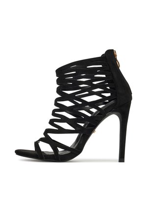 Czarna zamszowa sandałka na wysokim obcasie z krzyżującymi się paskami, odkrytymi palcami i zamkiem błyskawicznym z tyłu. Charakteryzuje się smukłym, spiczastym obcasem.