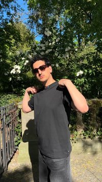 Jeune homme portant des lunettes de soleil et un t-shirt noir, posant à l'extérieur près d'un arbre en fleurs et d'une clôture en fer sous un soleil éclatant.