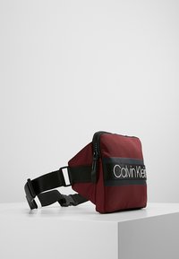 Rode nylon heuptas met zwarte strap en opvallende witte Calvin Klein branding. Vierkante vorm, ritsluiting, lichtgewicht textuur.