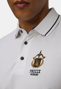 Polo bianco con colletto a punta, caratterizzato da strisce navy, tre bottoni e un'emblema ricamato con accenti dorati e testo.