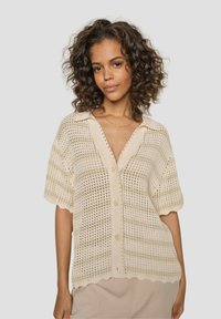 Light beige, short-sleeve knitted cardigan with a button-up front. Features a scalloped hem and textured open weave design.
