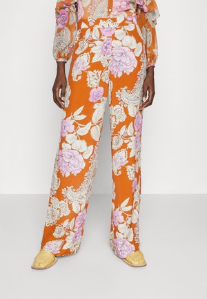 Szerokie pomarańczowe spodnie z dużymi kremowymi i różowymi wzorami kwiatowymi oraz paisley, noszone z beżowymi spiczastymi butami.