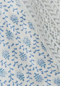 Tela de encaje blanca con bordado floral que cubre una tela blanca impresa con pequeños patrones azules de flores y ramas.