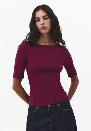 Kvinde med langt mørkt bølget hår iført en tætsiddende mørkelilla top med albuelange ærmer og mørkeblå jeans, stående mod en ensfarvet baggrund.