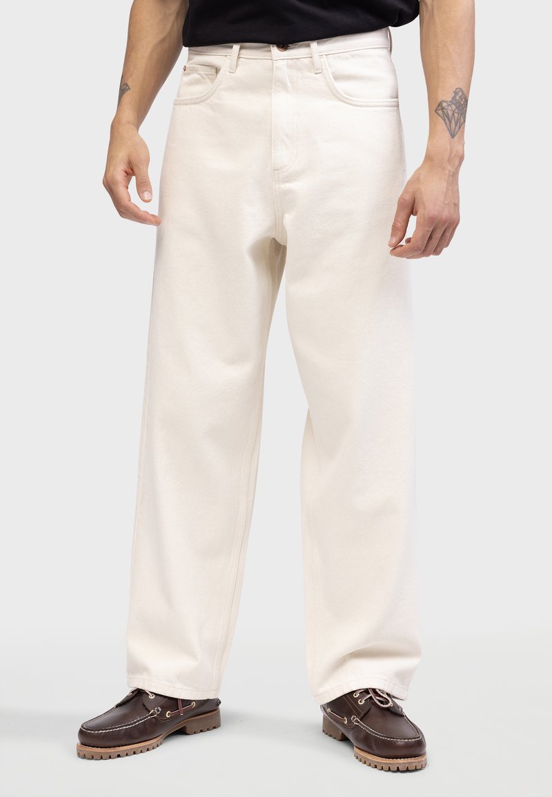 Jeans larges couleur crème en tissu épais avec une texture lisse. Comprend une fermeture à bouton et des poches avant, portés avec des chaussures foncées.