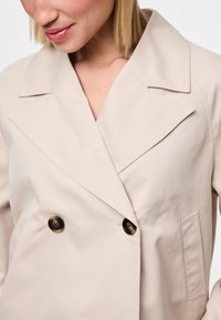 Donna che indossa un cappotto beige doppiopetto con grandi rever e bottoni scuri, mostrata dalla collottola fino a metà torso.
