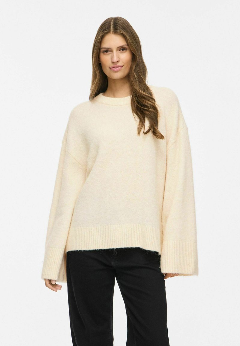 Femme aux longs cheveux bruns portant un pull ample crème et un pantalon noir, debout devant un fond blanc, regardant droit devant.