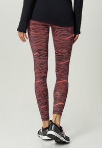 Mönstrade leggings i flera färger har ett vågigt mönster i rött och mörka nyanser. Tyget är stretchigt och figurnära, tillsammans med svarta träningsskor.