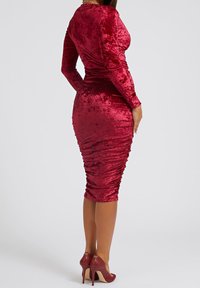 Robe midi en velours rouge à manches longues avec des détails froncés le long du dos. Associée à des chaussures à talons assorties. Texture lisse.